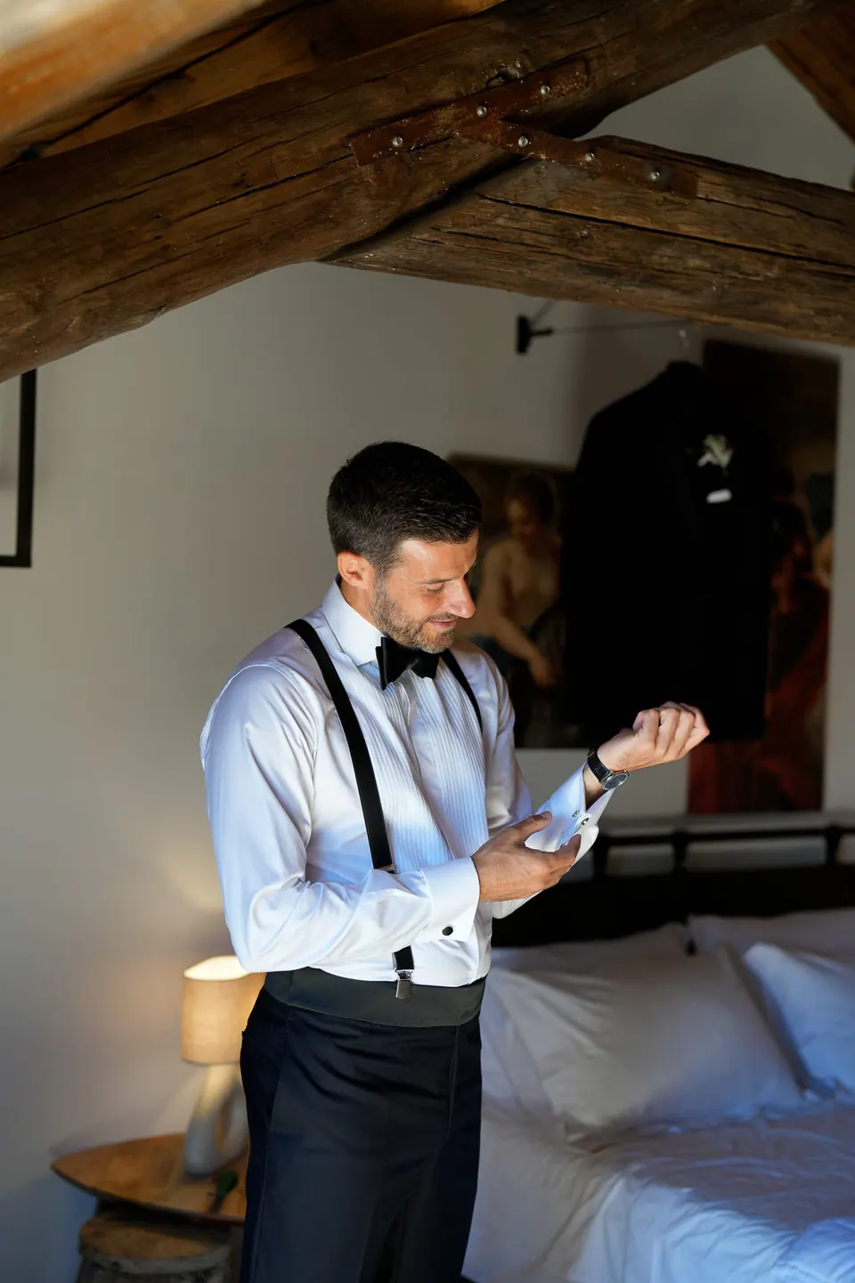 Lo sposo allaccia i gemelli accanto al letto, bretelle sulla camicia bianca.