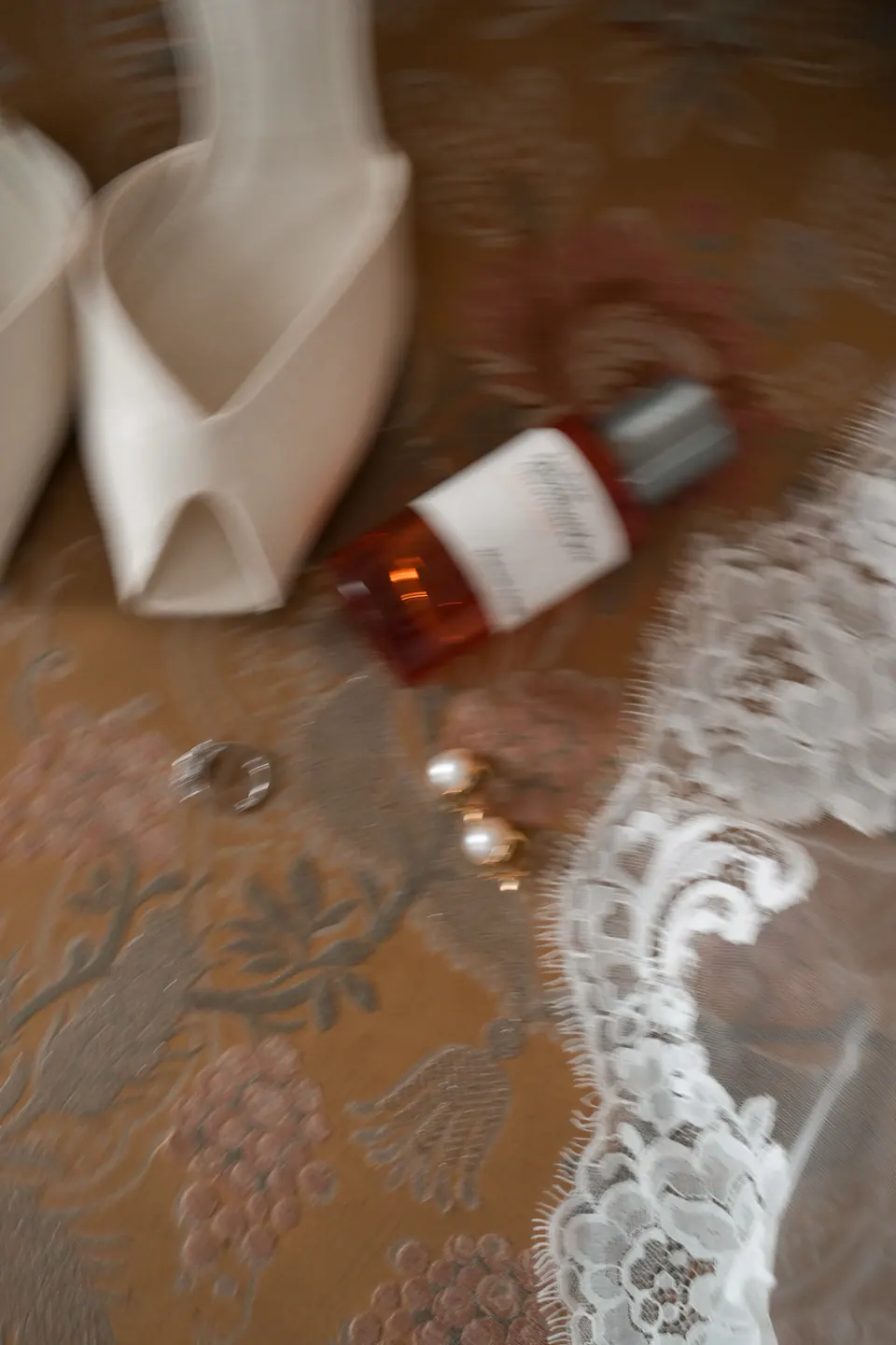Scarpe da sposa, profumo e orecchini di perle su una poltrona in broccato accanto al pizzo del velo.