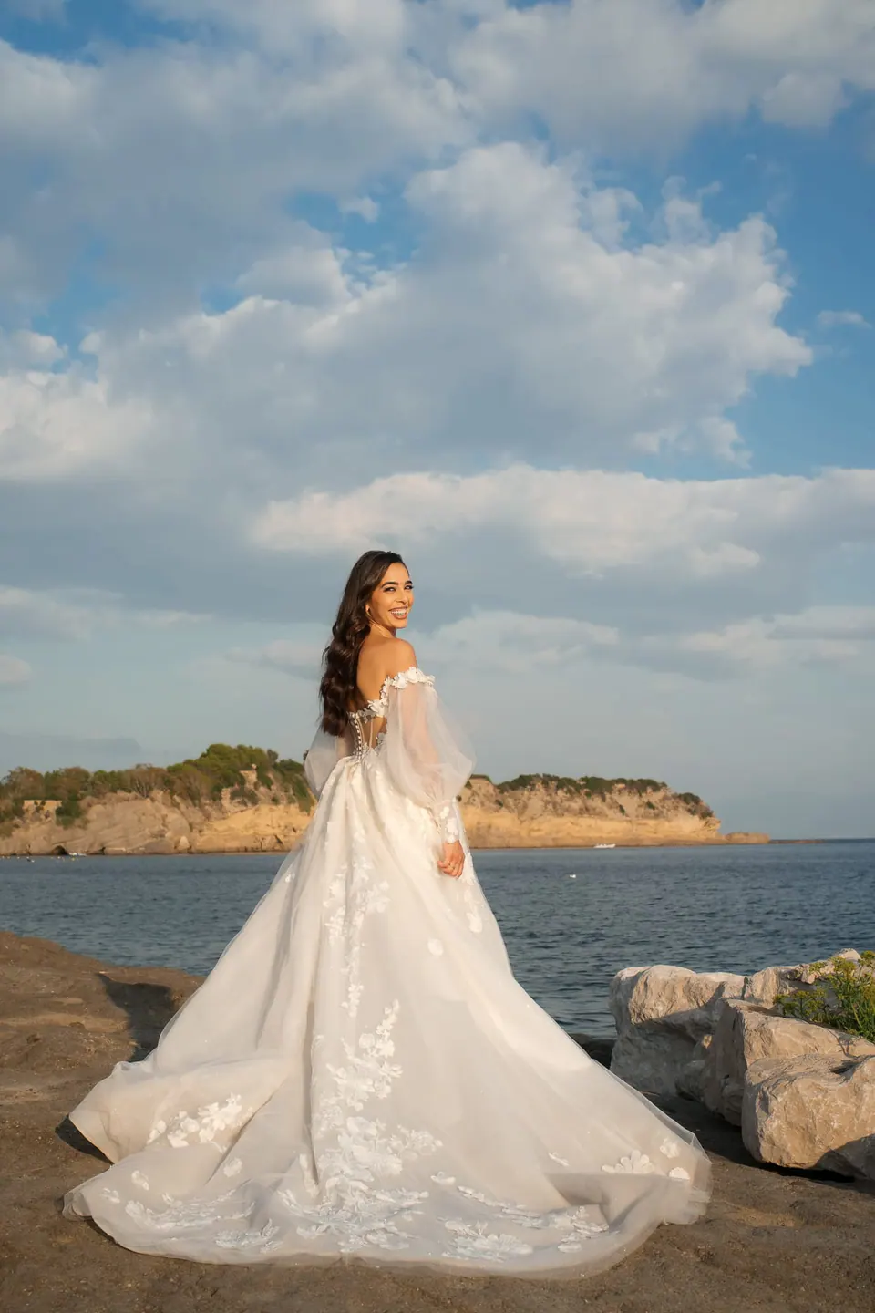 Bride standing on the rocks by the sea, a green headland behind her.