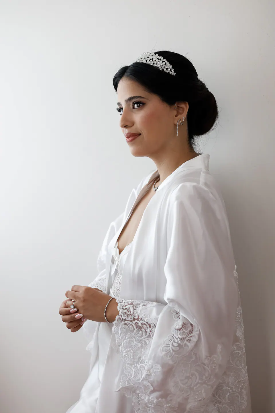 La mariée près de la fenêtre dans son peignoir aux poignets de dentelle, les cheveux relevés sous le diadème.