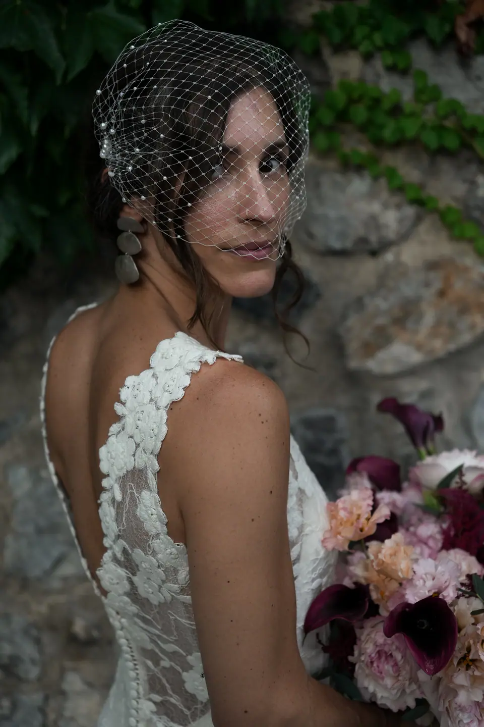 Bride in a birdcage veil looking over her shoulder, her bouquet of pink flowers.