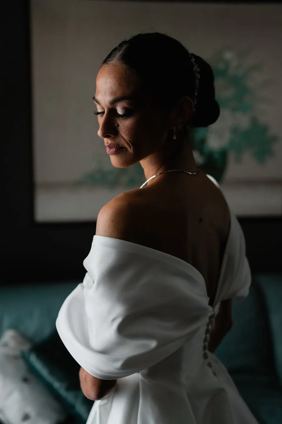 Sposa che guarda in basso sopra la spalla con l'abito a spalle scoperte, luce laterale morbida.