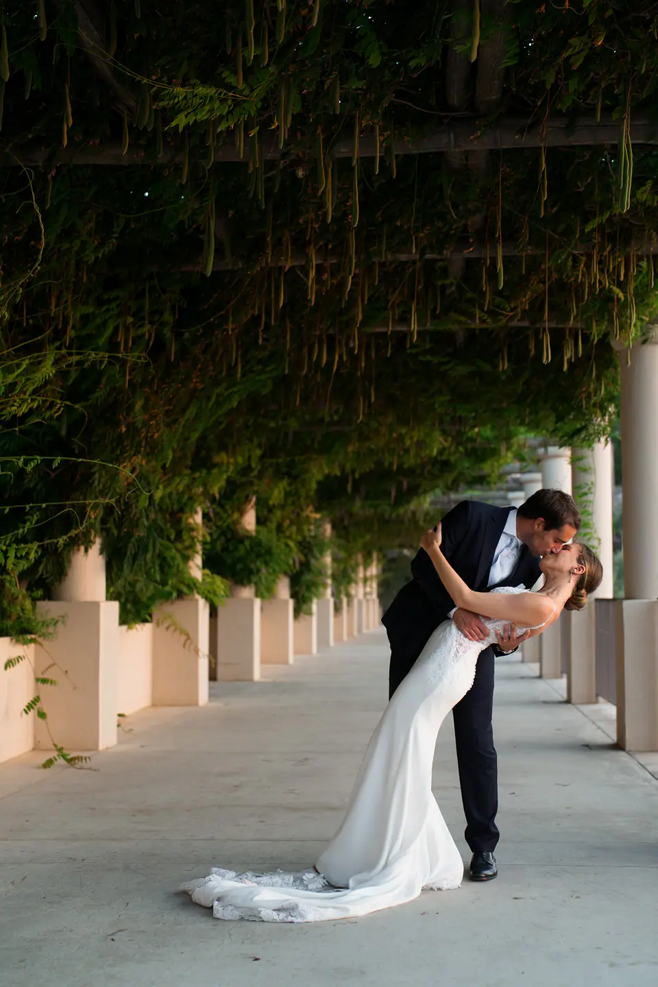 Groom dipping the bride under a colonnade covered in green.