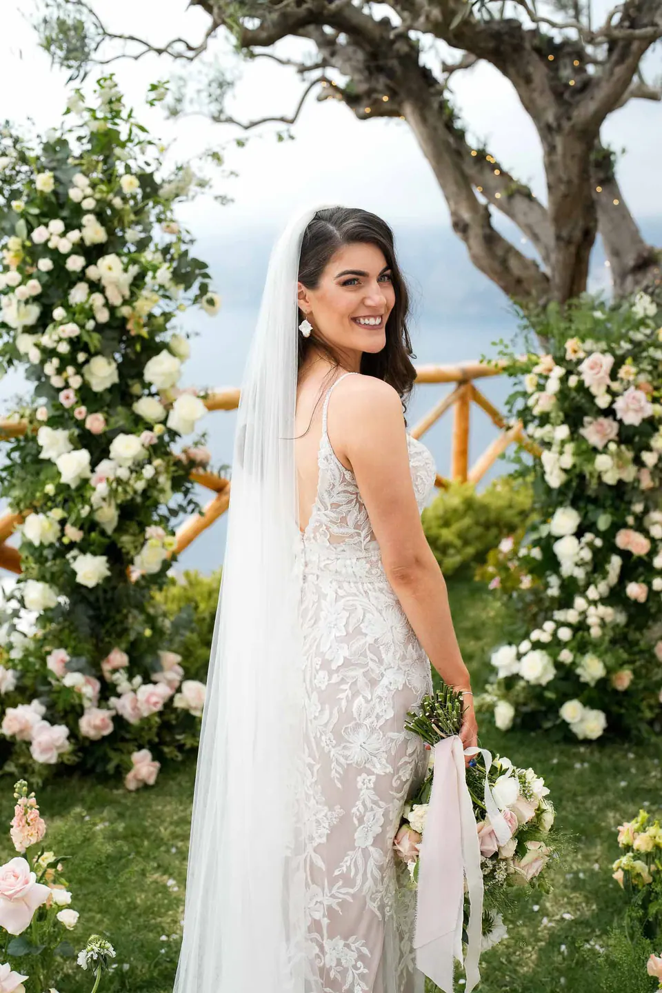 La sposa sotto un arco floreale con vista sul mare, la luce del sole a illuminarne l'abito.