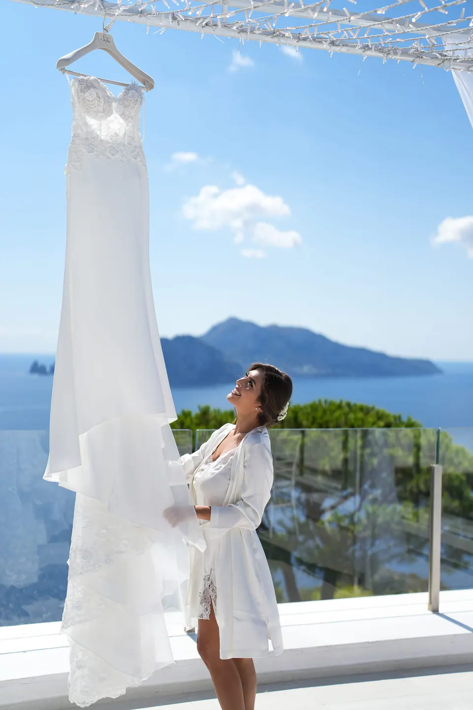 Sposa in vestaglia accanto all'abito appeso al pergolato, Capri all'orizzonte.