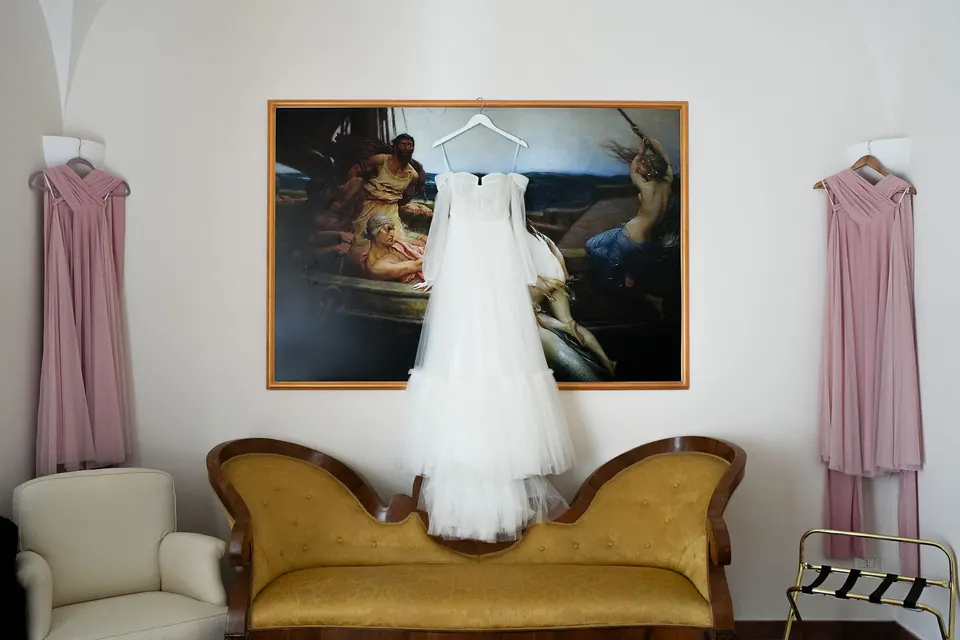 Das Brautkleid über einem goldenen Sofa zwischen zwei rosa Brautjungfernkleidern, unter einem klassischen Gemälde.