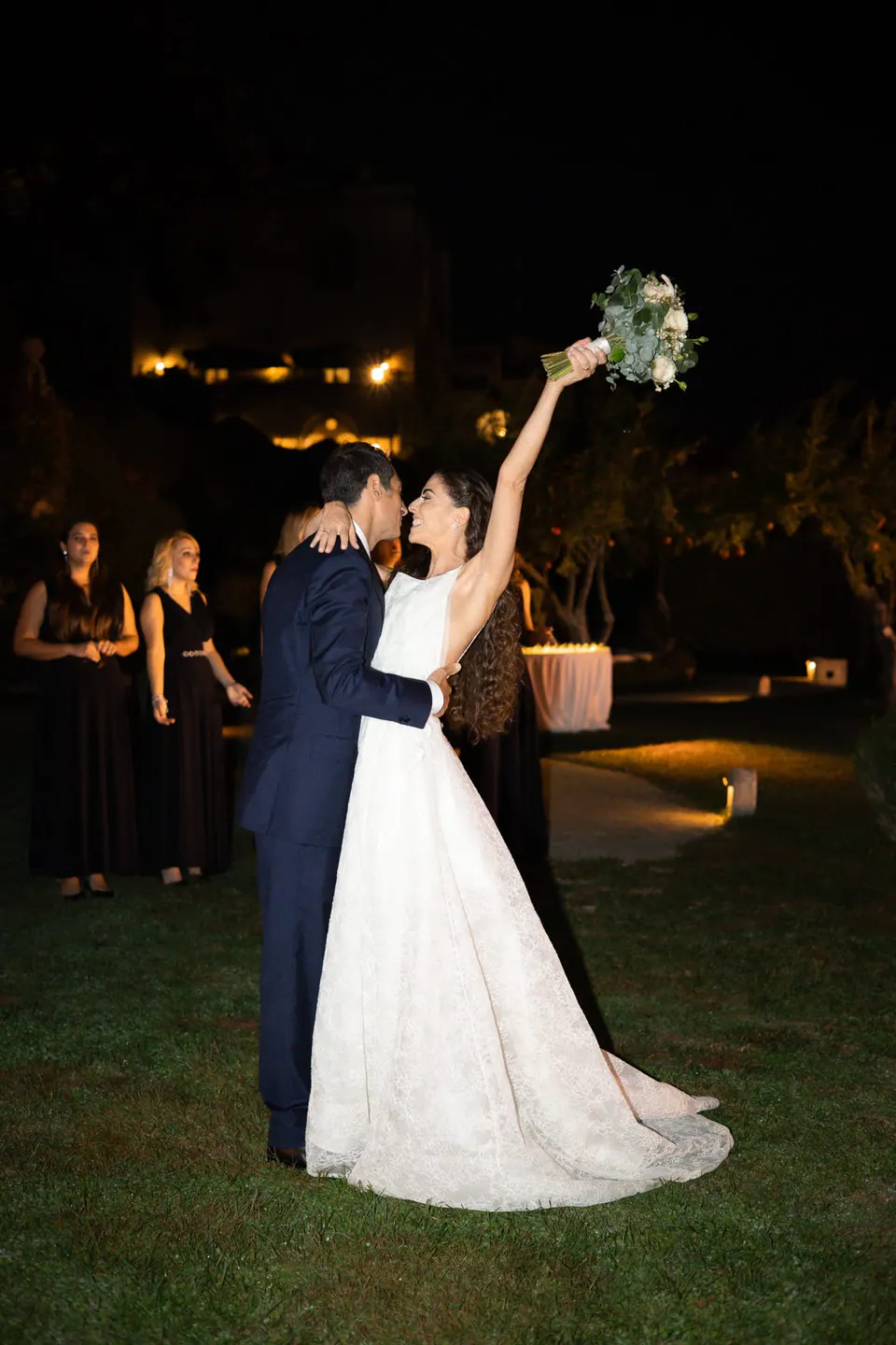 Bride raising her bouquet on the lawn at night as the groom kisses her.