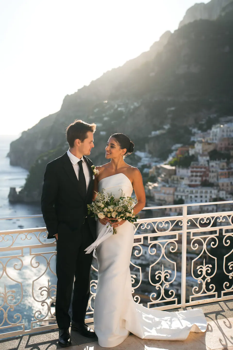 Bride and groom at the railing, bouquet in hand, Positano behind them.