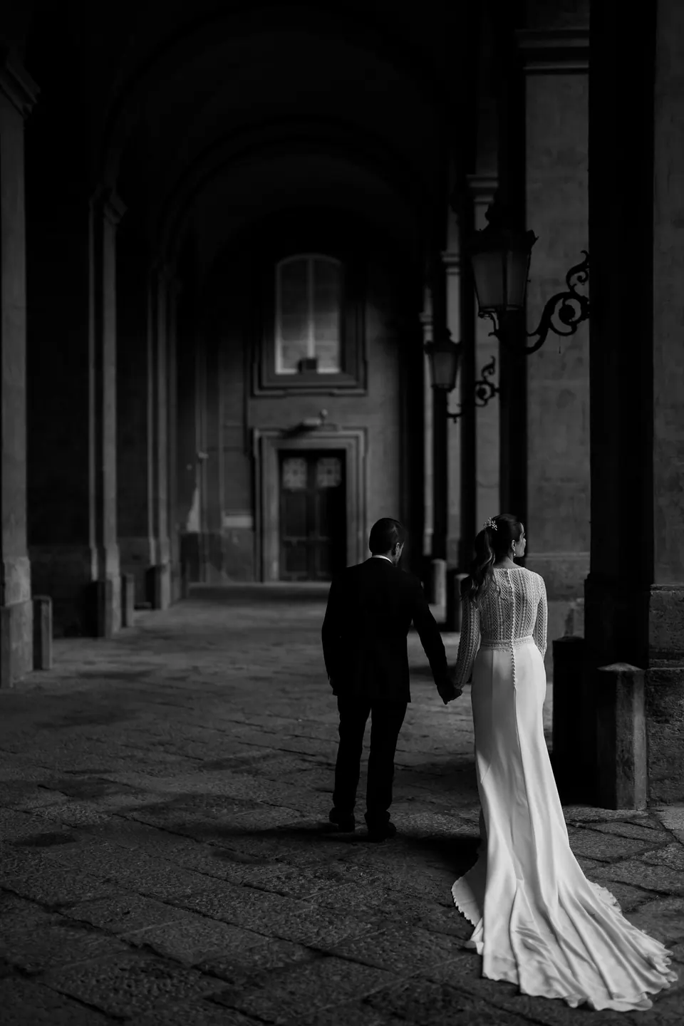 Black and white photo of the couple beneath a dark arcade of the palace, her train on the stone.
