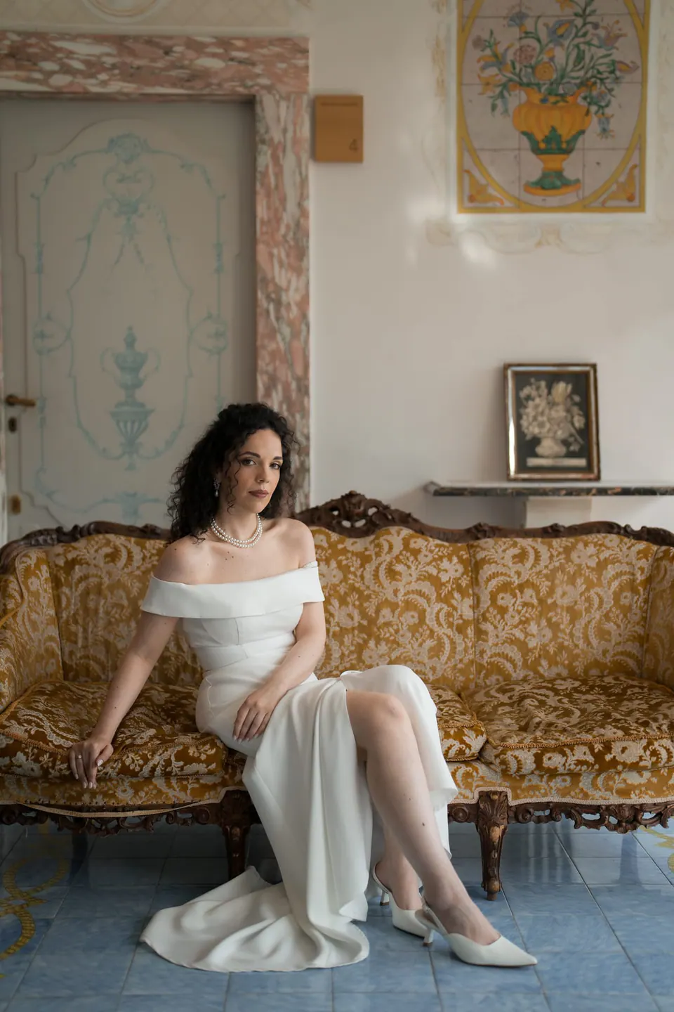 La novia sentada en un sofá antiguo bajo cuadros enmarcados, con luz suave de ventana en Villa Ortensia D’Avalos.