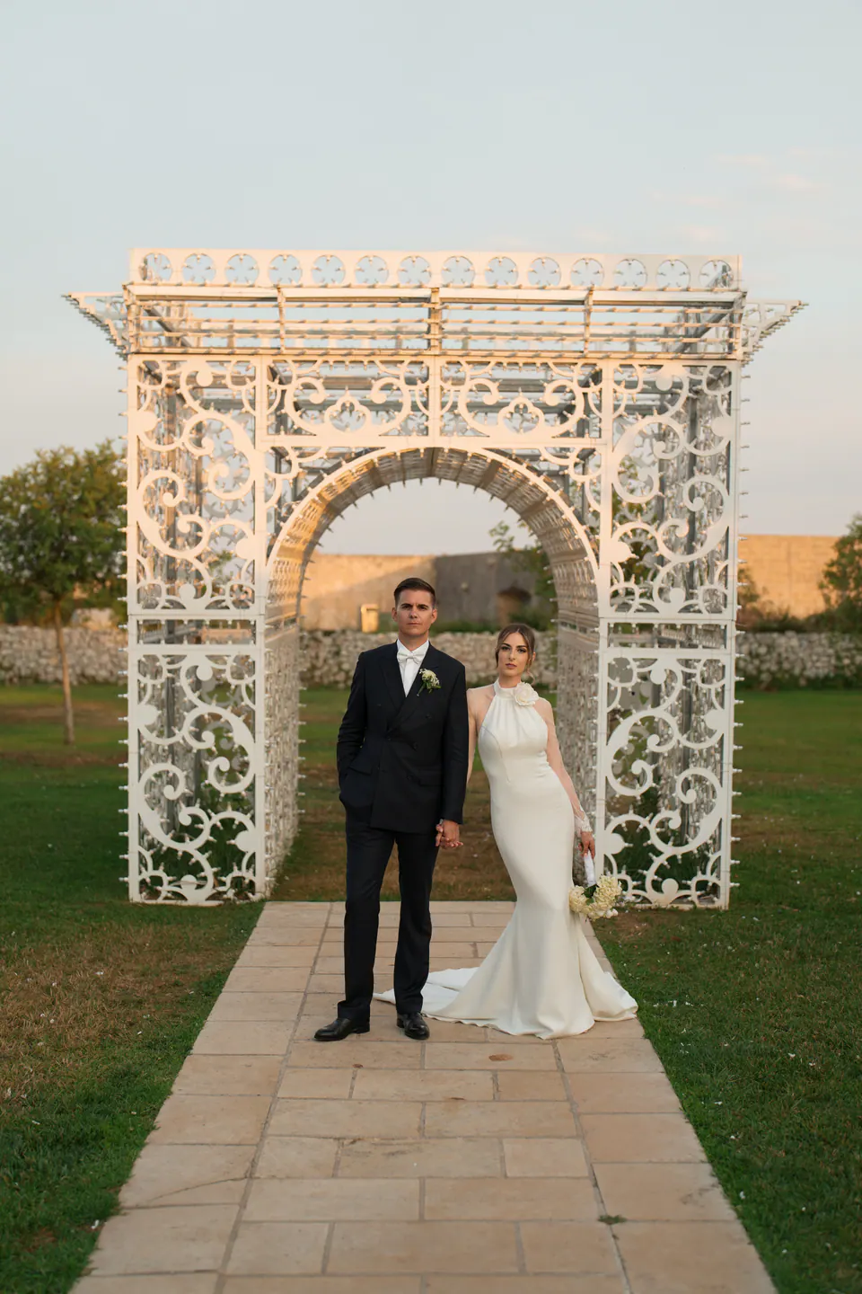 Gli sposi sotto un arco bianco in ferro battuto nel giardino di Tenuta Sant'Elia.