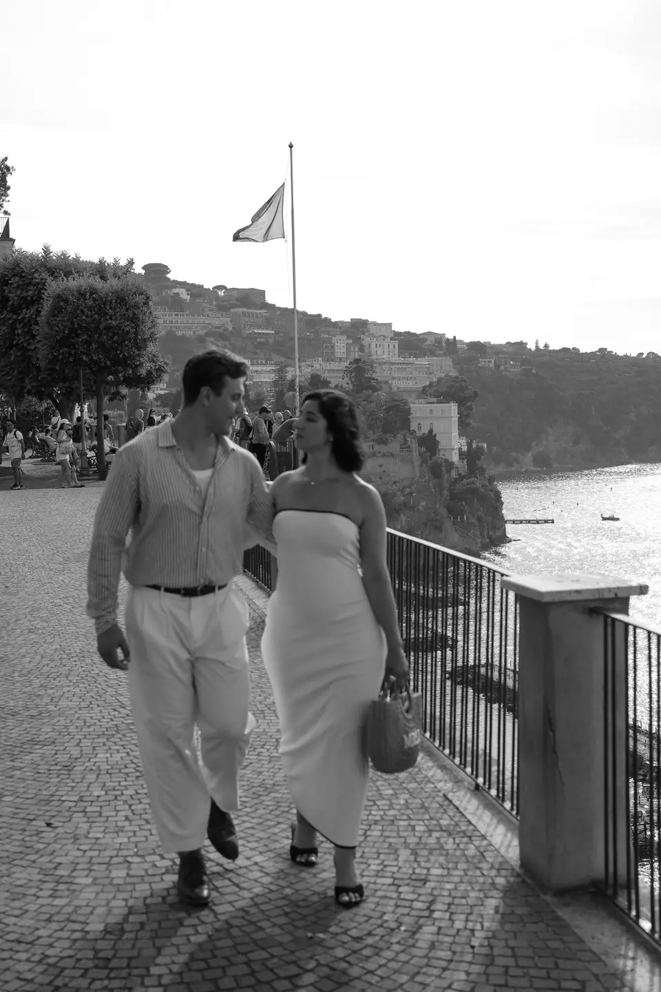 Black and white photo of the couple walking on a terrace above the coast.