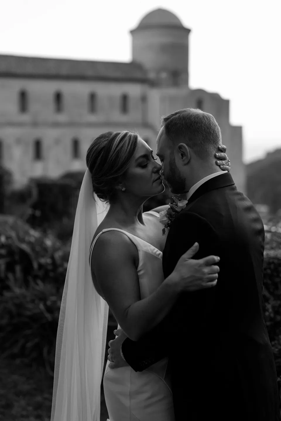 Black and white photo of bride and groom kissing, a bell tower of Ravello behind them