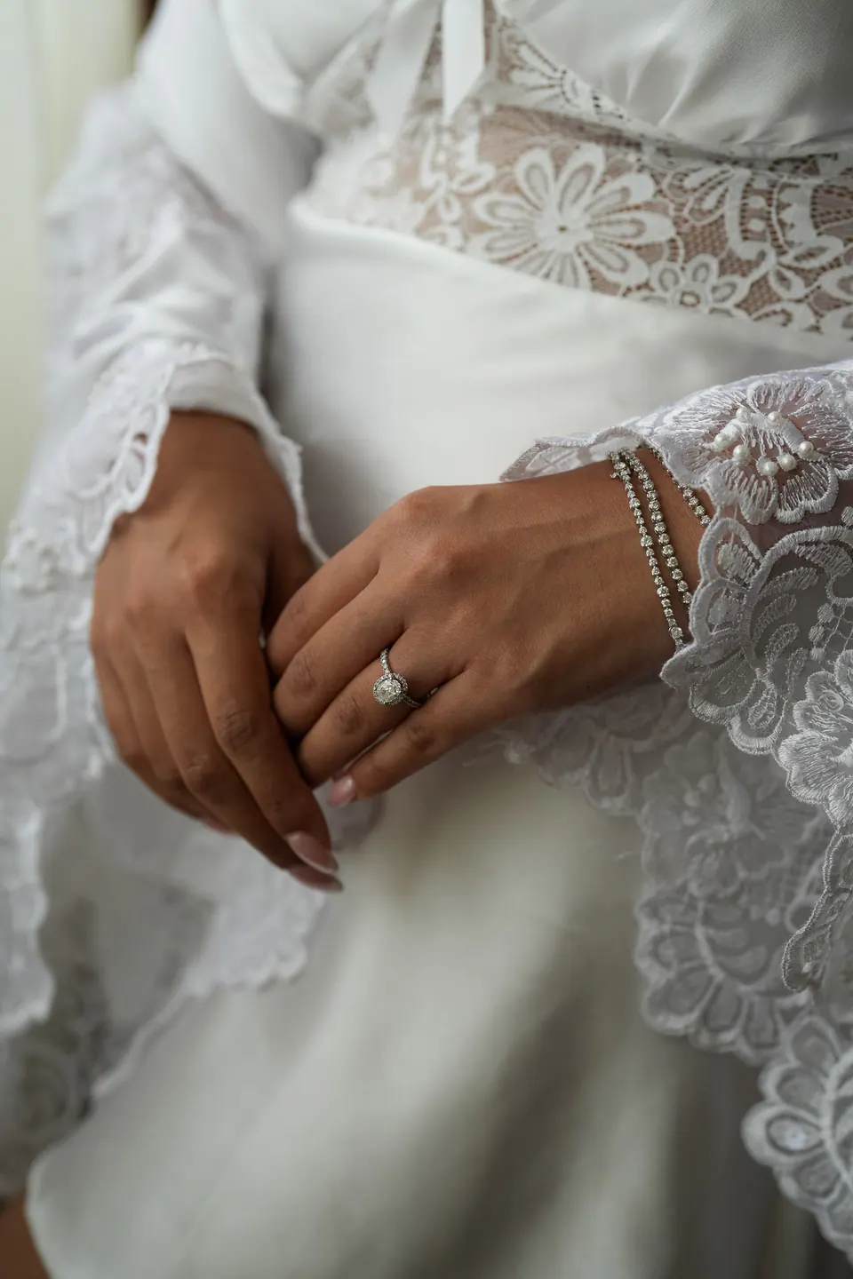 Détail des mains de la mariée, la bague et le bracelet de perles sur la dentelle du peignoir.