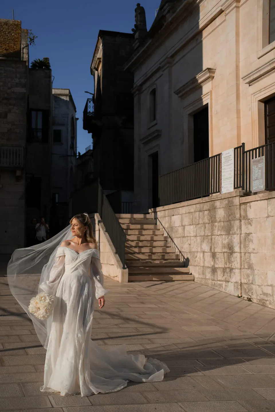 La novia en una calle al anochecer, con el velo levantado por el viento.