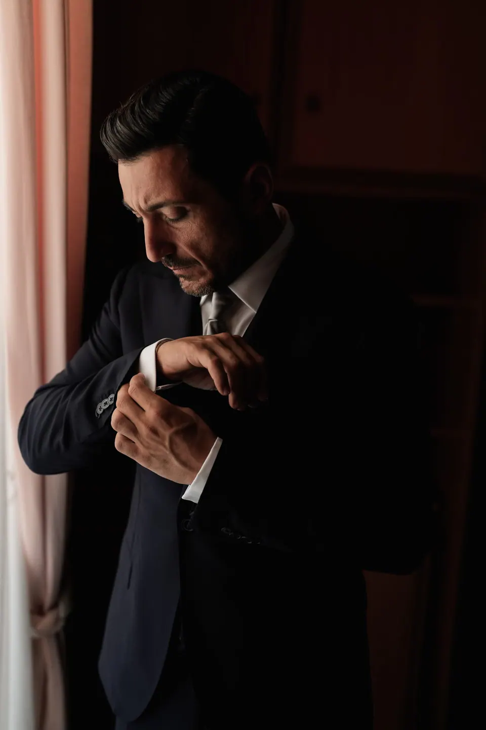 Groom fastening his cufflinks by a curtain in soft light.