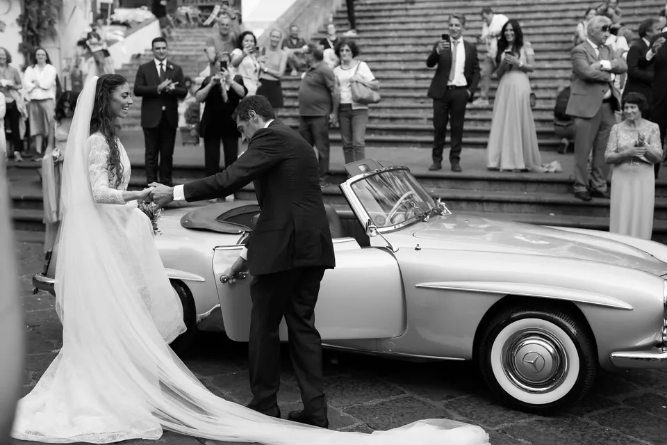 Black and white photo of the groom helping the bride into the vintage convertible below the cathedral steps.