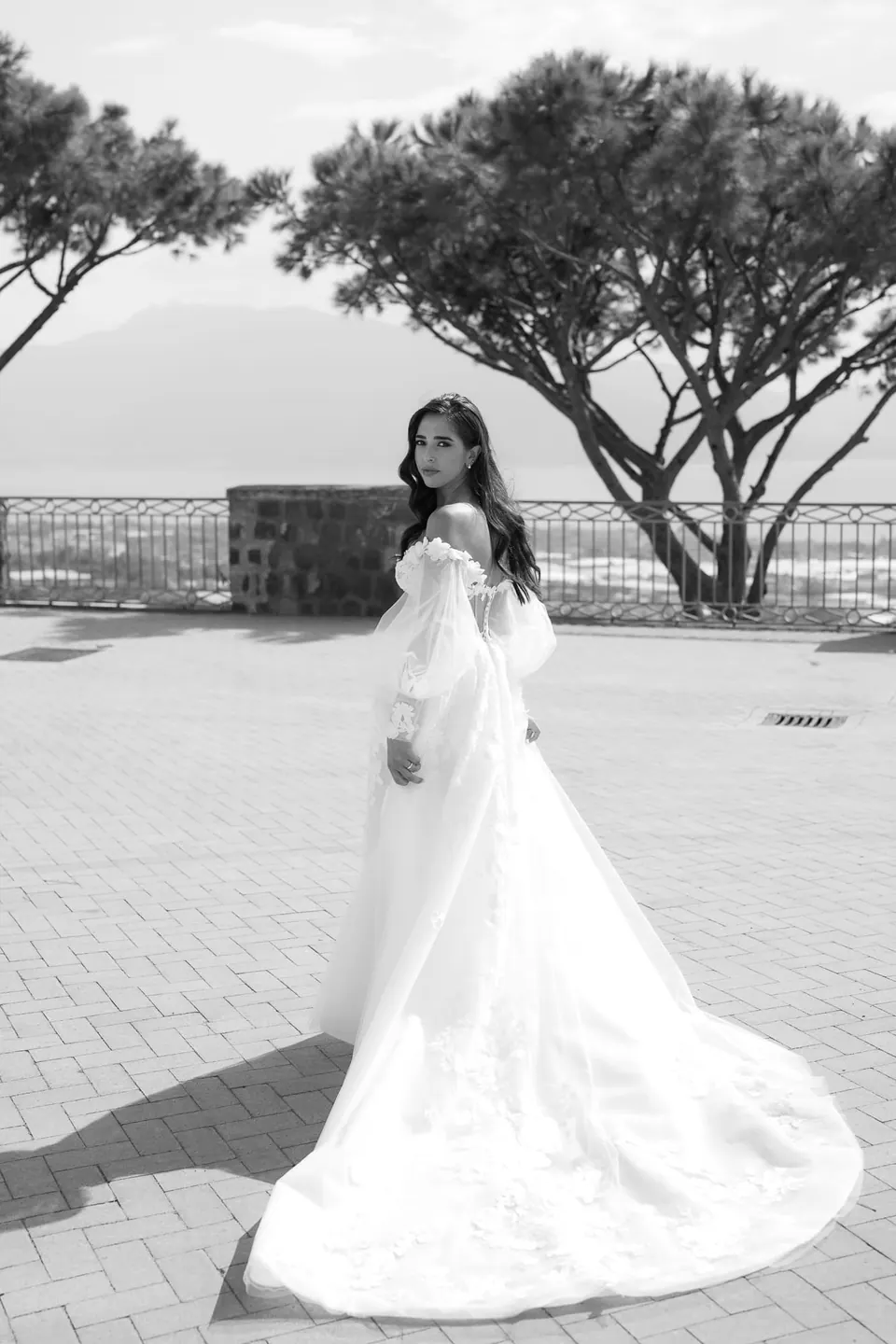 Black and white photo of the bride on the terrace beneath umbrella pines, her long train spread behind her.