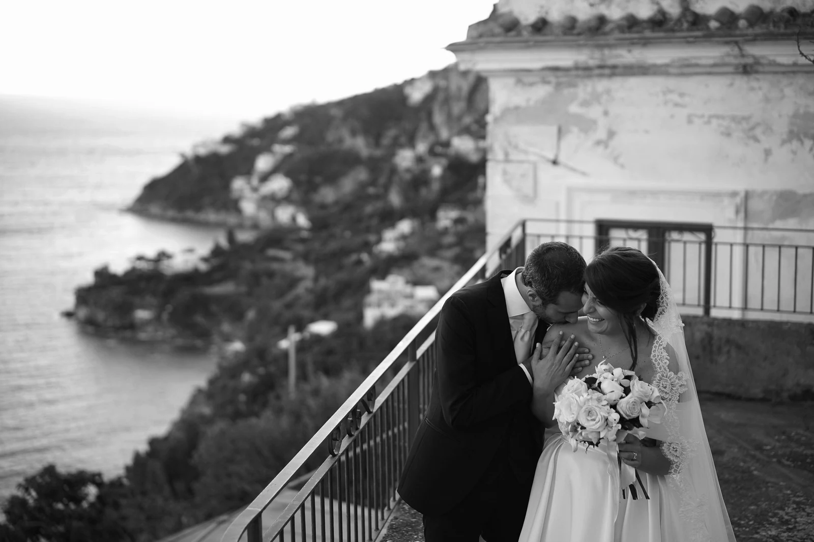 Couple sharing a kiss overlooking the Amalfi coastline, waves glimmering below them.