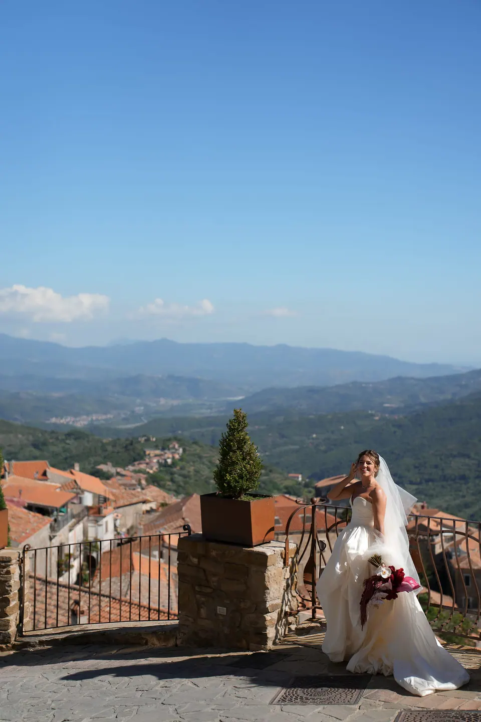 Sposa sulla terrazza del castello con il bouquet rosso, tetti e colline del Cilento alle spalle.