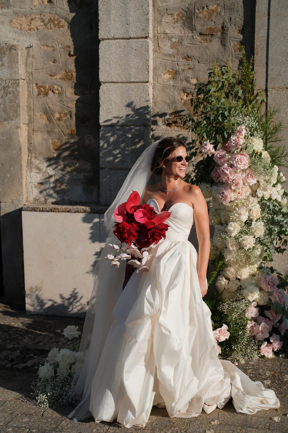 Sposa con occhiali da sole e rose rosse accanto a un arco fiorito sul muro del castello.