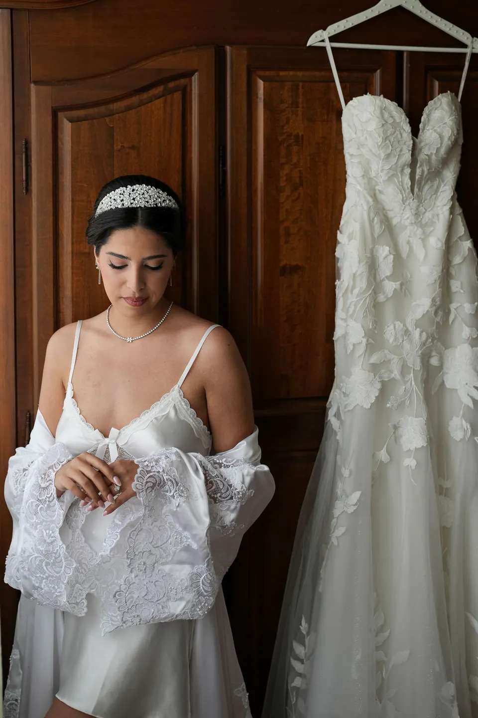 La mariée en peignoir près de l’armoire en bois où est suspendue la robe de mariée.