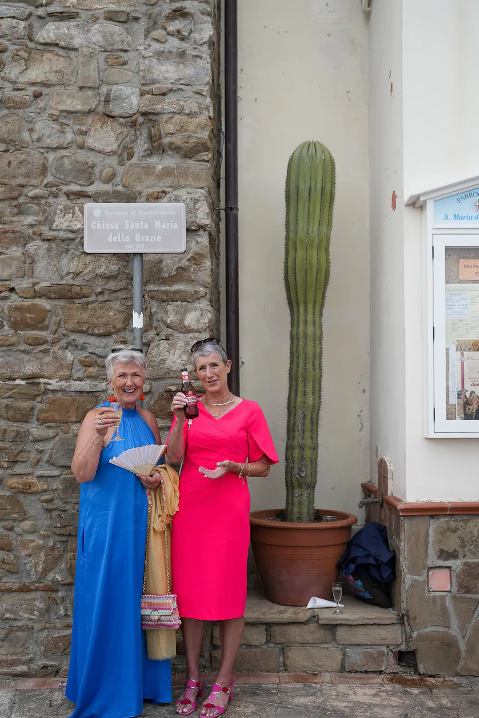 Zwei Gäste in leuchtend blauem und rosa Kleid an einer Steinmauer mit hohem Kaktus.