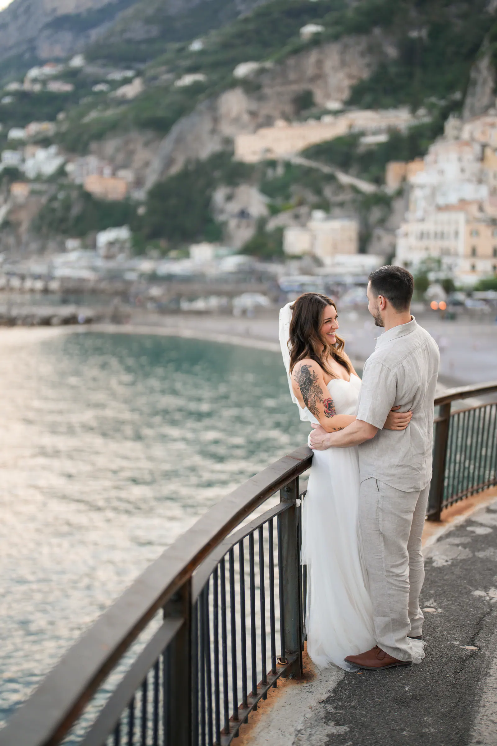 Couple embracing at a railing above the bay of Amalfi, the town and the mountains behind them.