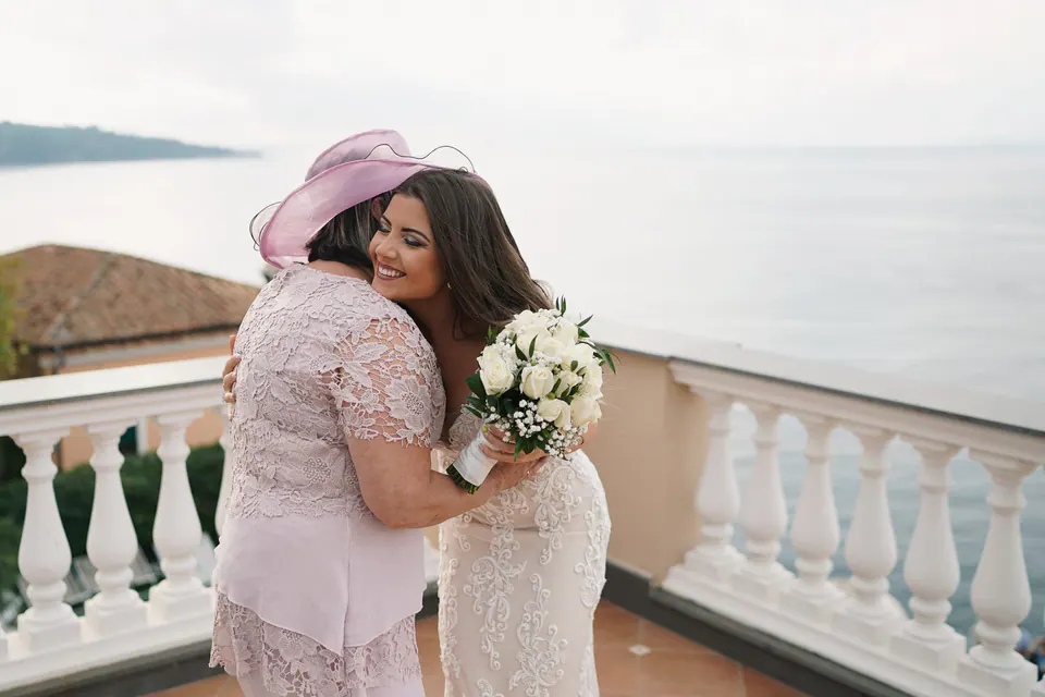 Bride hugging her mother in a pink hat on the terrace, bouquet in hand.
