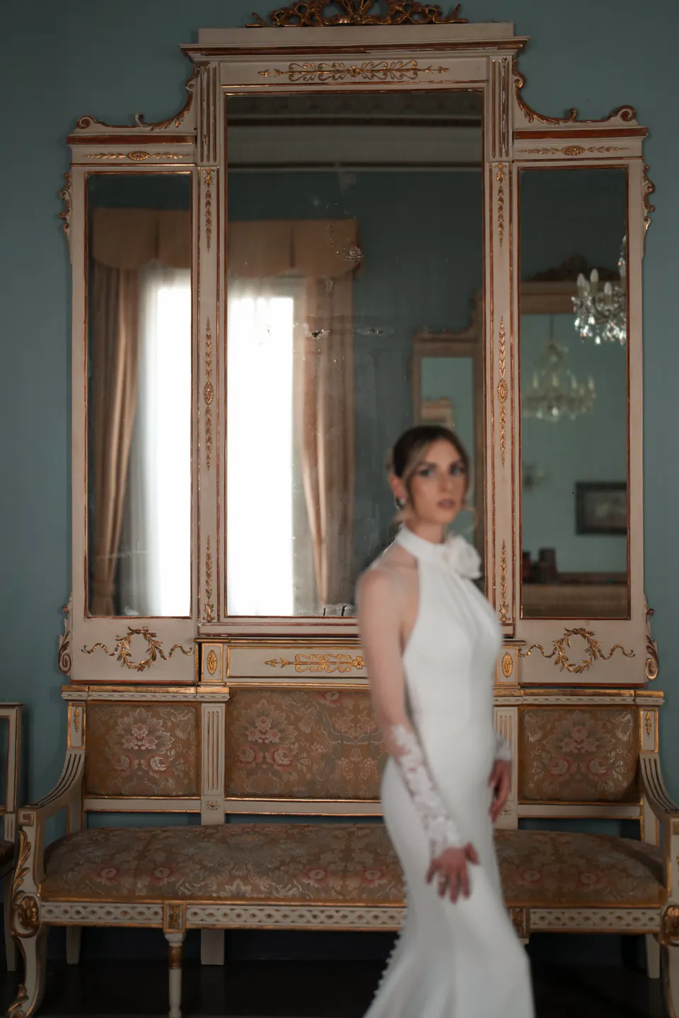 La sposa con l'abito all'americana davanti a una specchiera dorata in una stanza celeste.