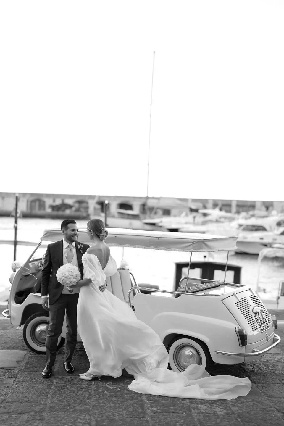 Schwarzweißfoto des Paares neben einem Oldtimer am Hafen.