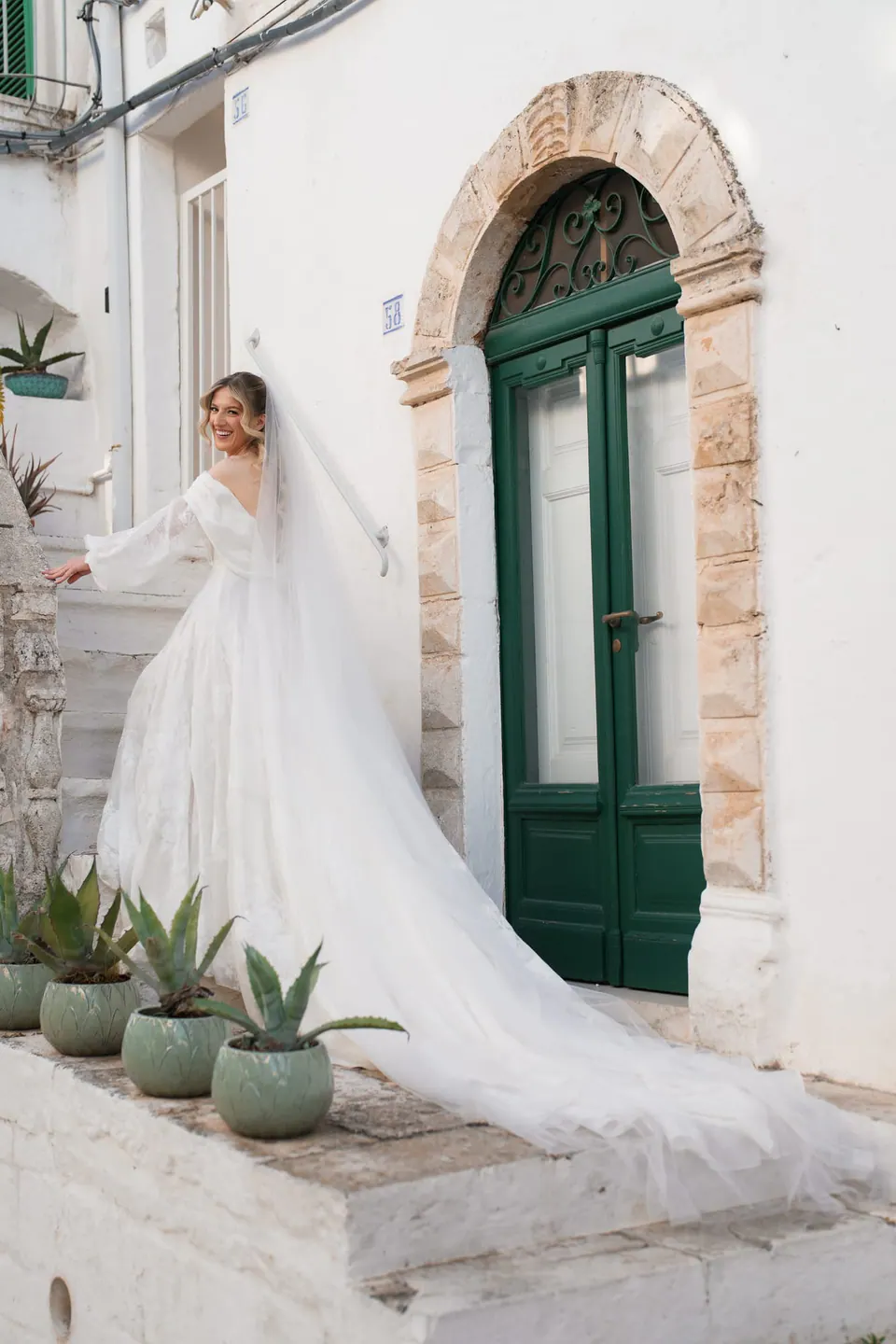 La novia en los escalones junto a una puerta verde en arco, con el velo extendido detrás.