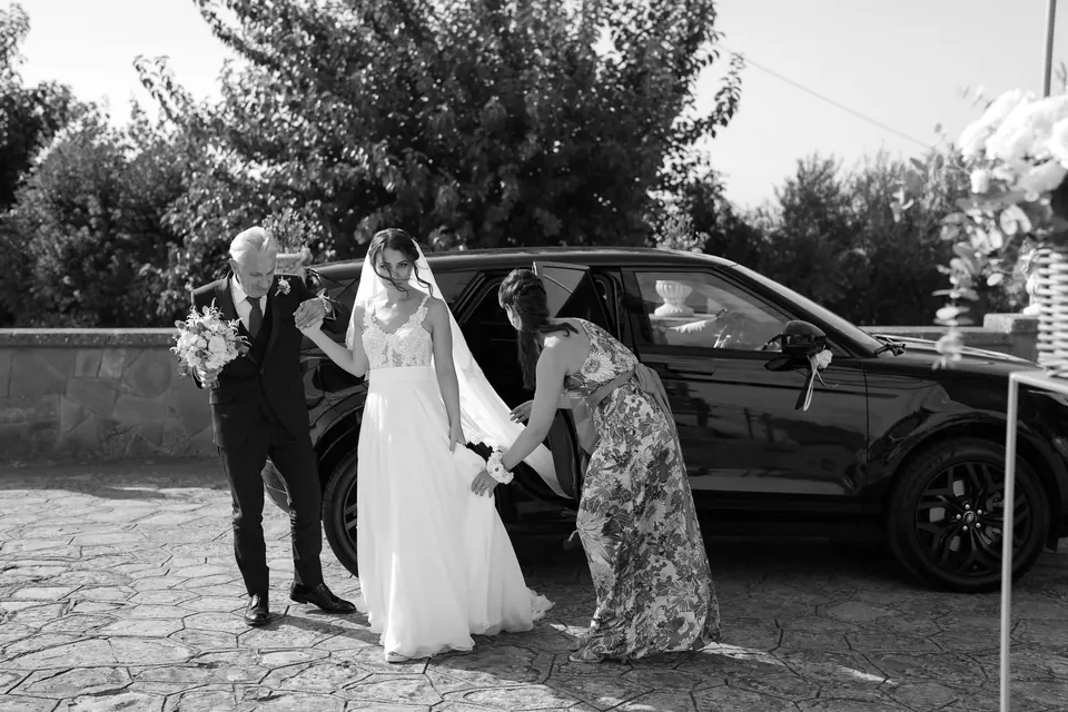 Black and white photo of the bride stepping out of the car with her father as her dress is arranged.