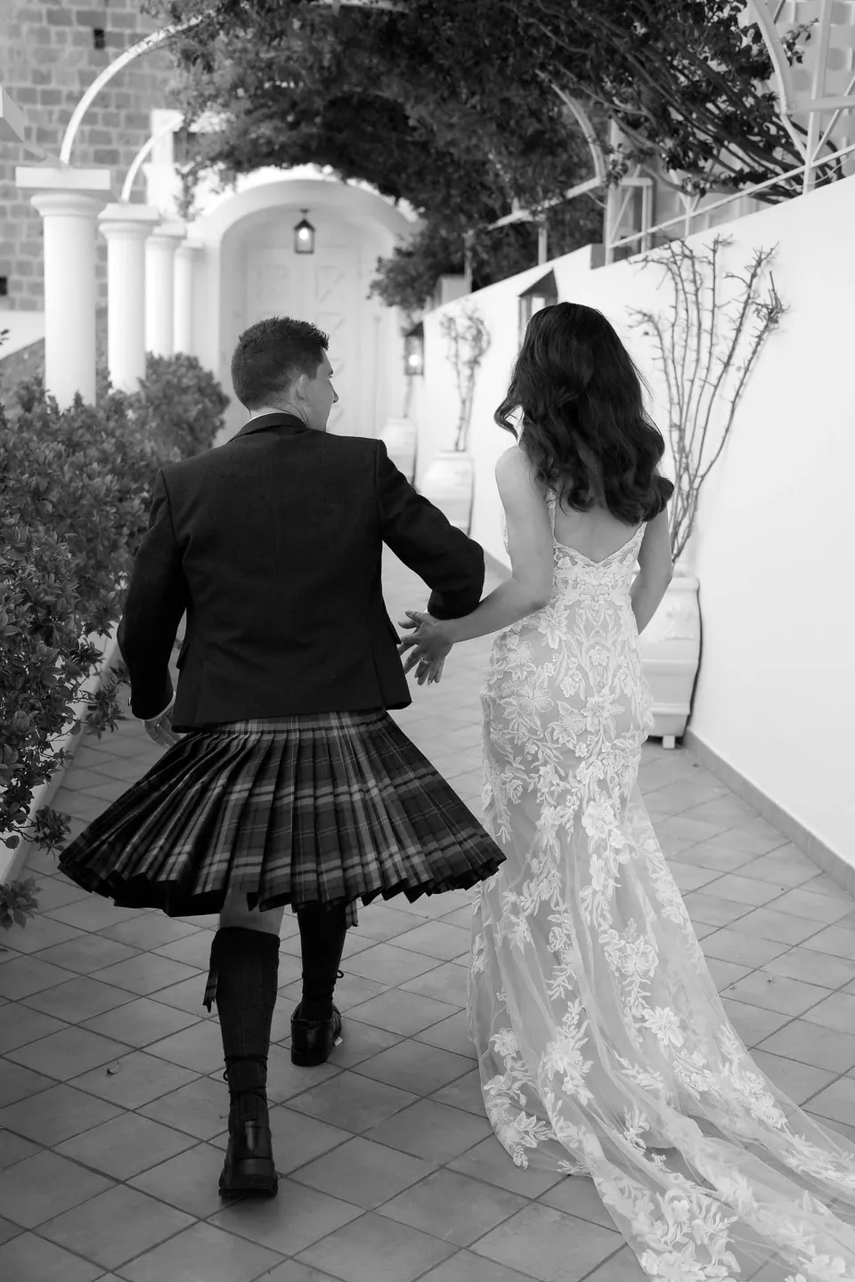 Foto in bianco e nero dello sposo in kilt che cammina accanto alla sposa lungo la terrazza.