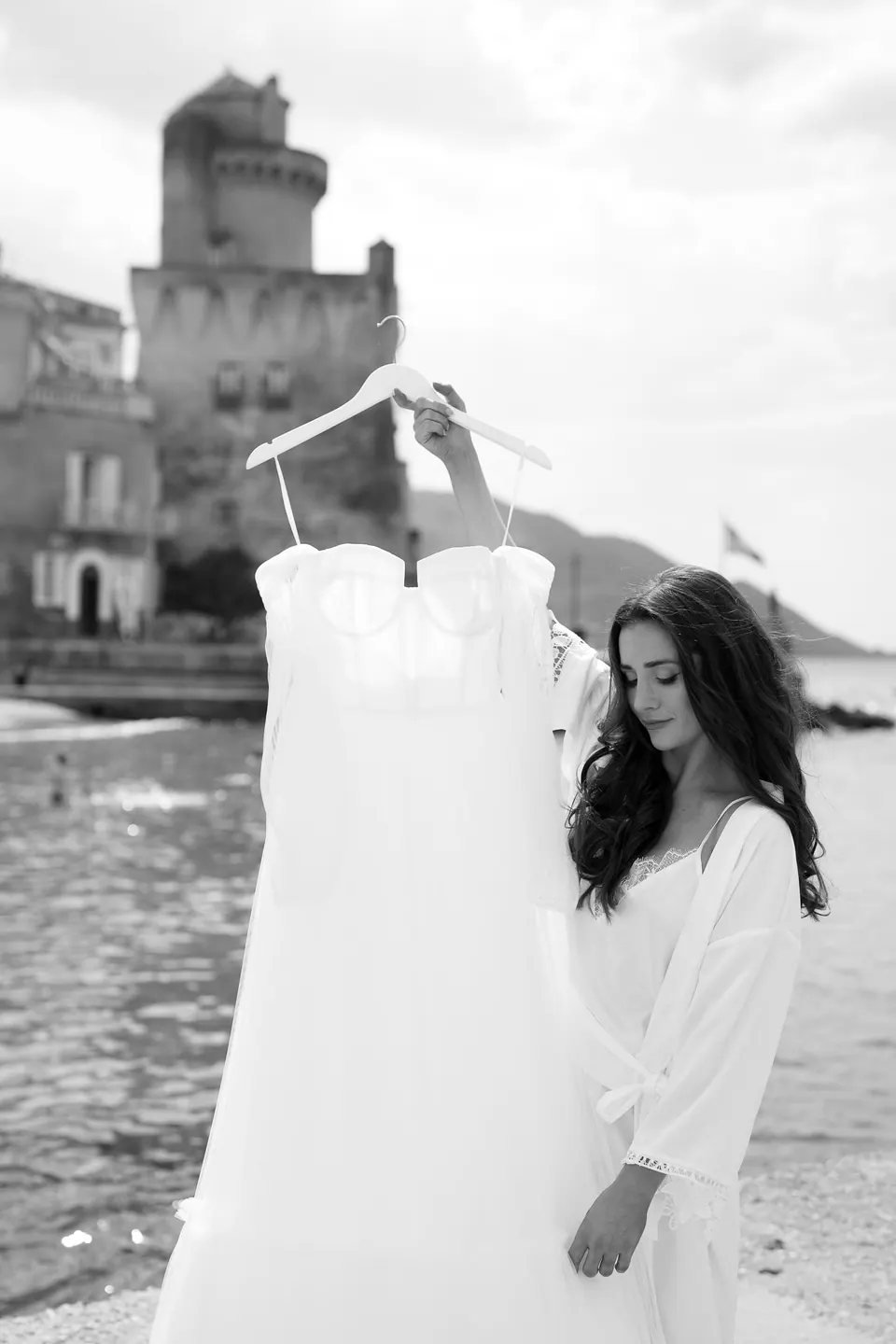 Schwarzweißfoto: Die Braut hält ihr Kleid am Bügel am Meer, im Hintergrund der Turm.