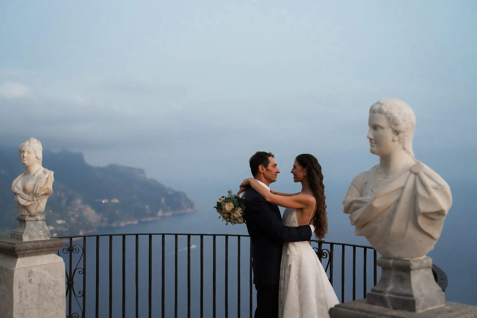 Sposi abbracciati accanto a un busto in marmo sulla Terrazza dell'Infinito, la costa alle spalle.