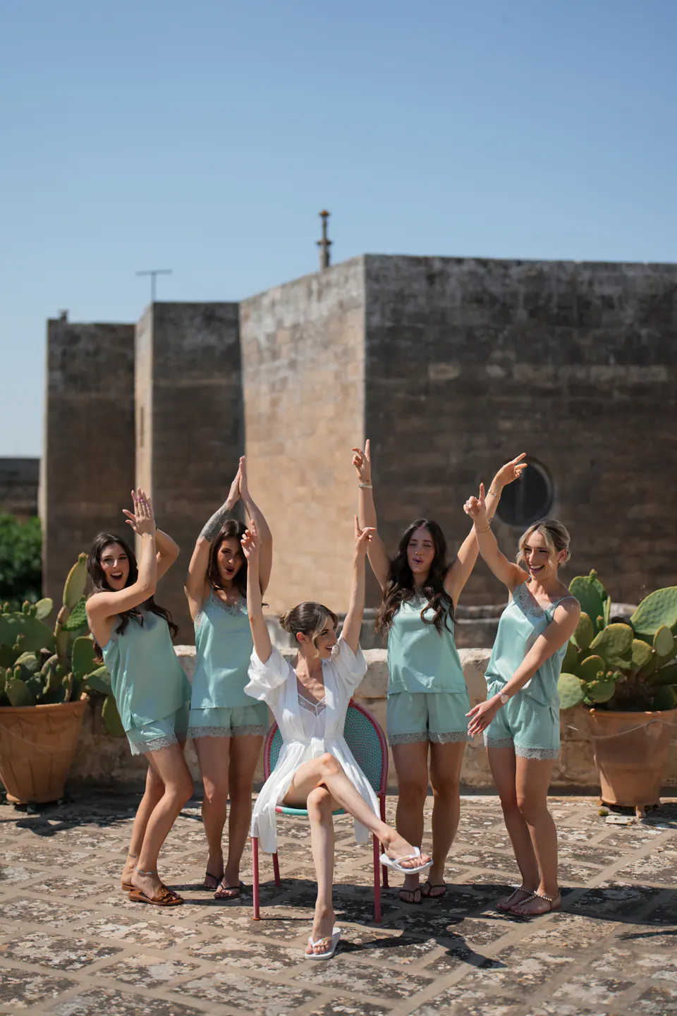 Le damigelle che esultano intorno alla sposa su una terrazza in pietra con antiche torri e fichi d'India.