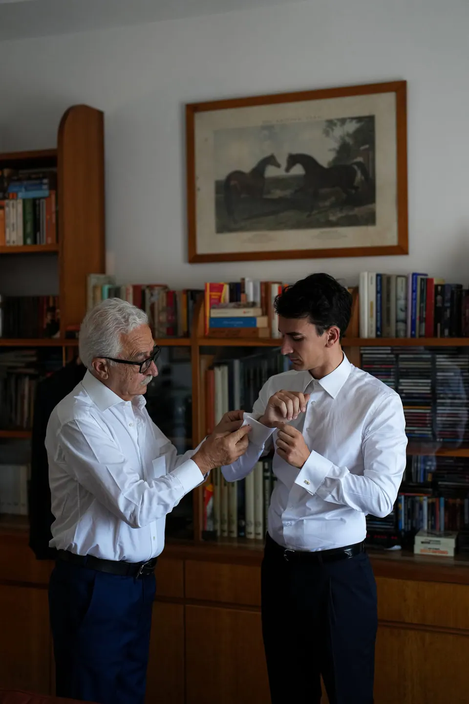 An older man helping the groom with his cufflinks in a book-lined room