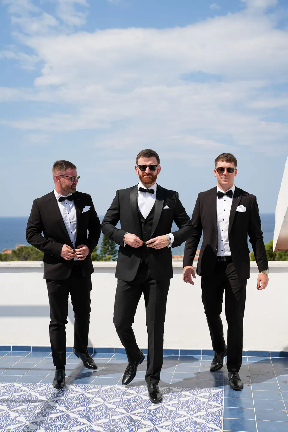 Groom and groomsmen in tuxedos walking across a majolica terrace with the sea behind them.