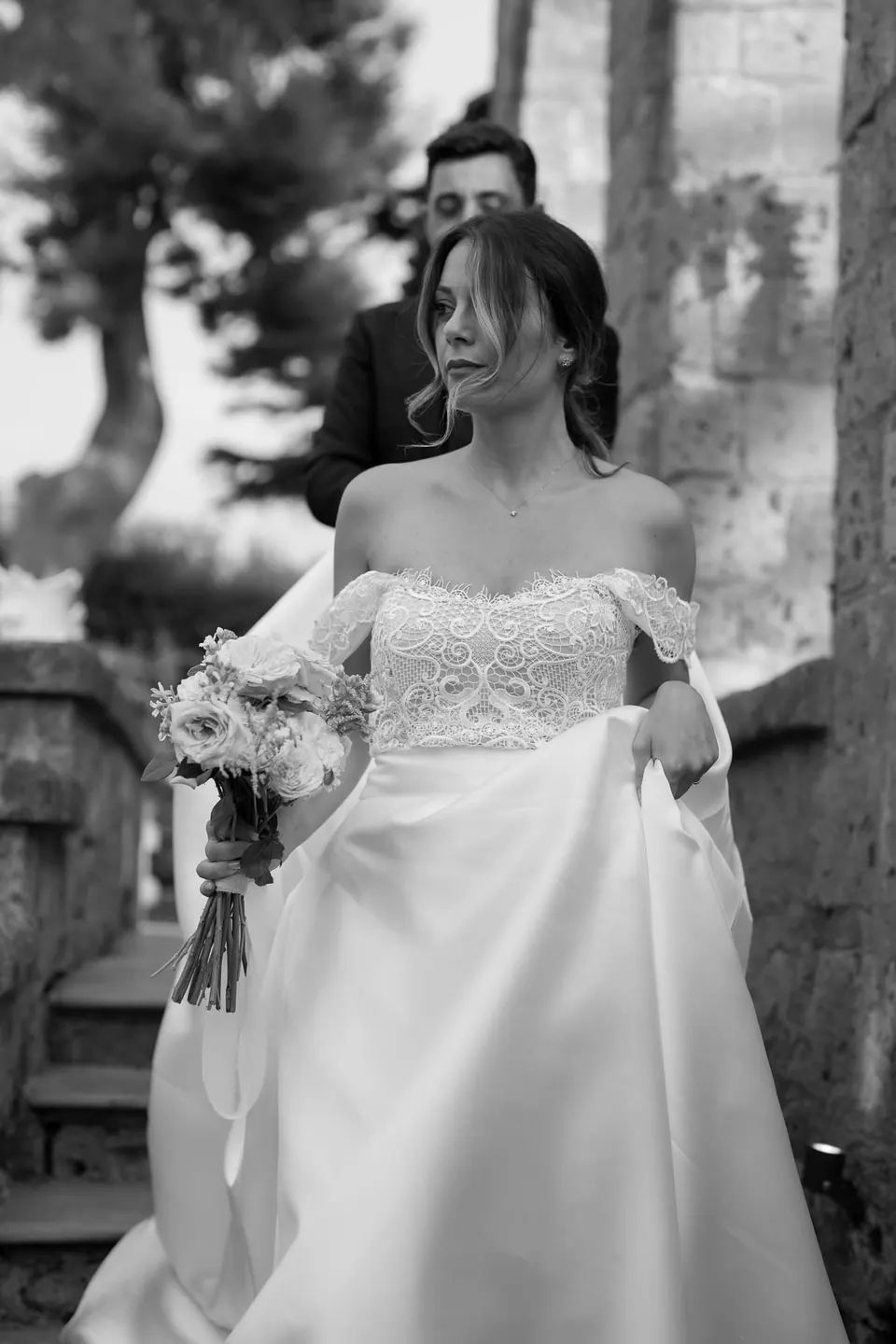 Black and white photo of the bride walking in the garden with the groom behind her