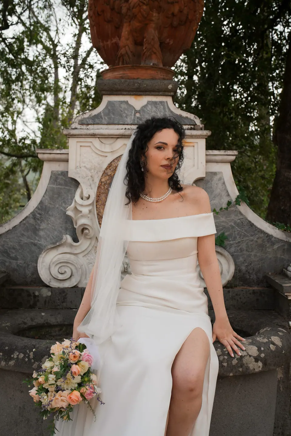 La novia sentada en la fuente con su ramo, la abertura del vestido y el velo alrededor.