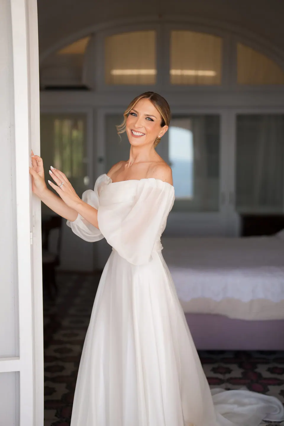 Die Braut lächelt im schulterfreien Kleid am Fenster.