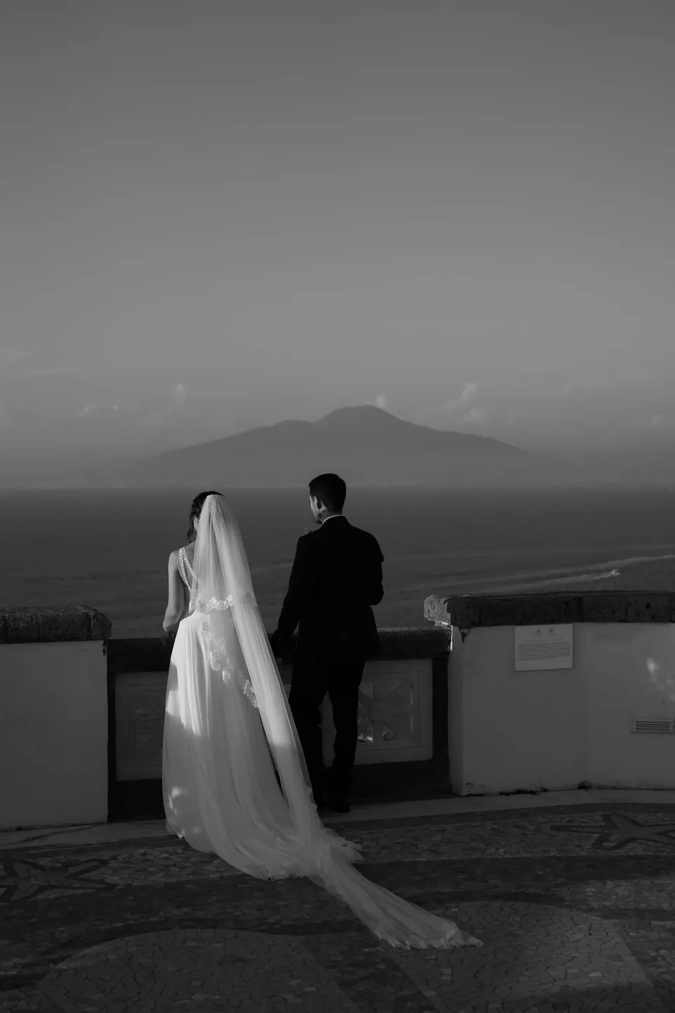 Black and white photo of the couple from behind on the terrace, Vesuvius across the Gulf of Naples.