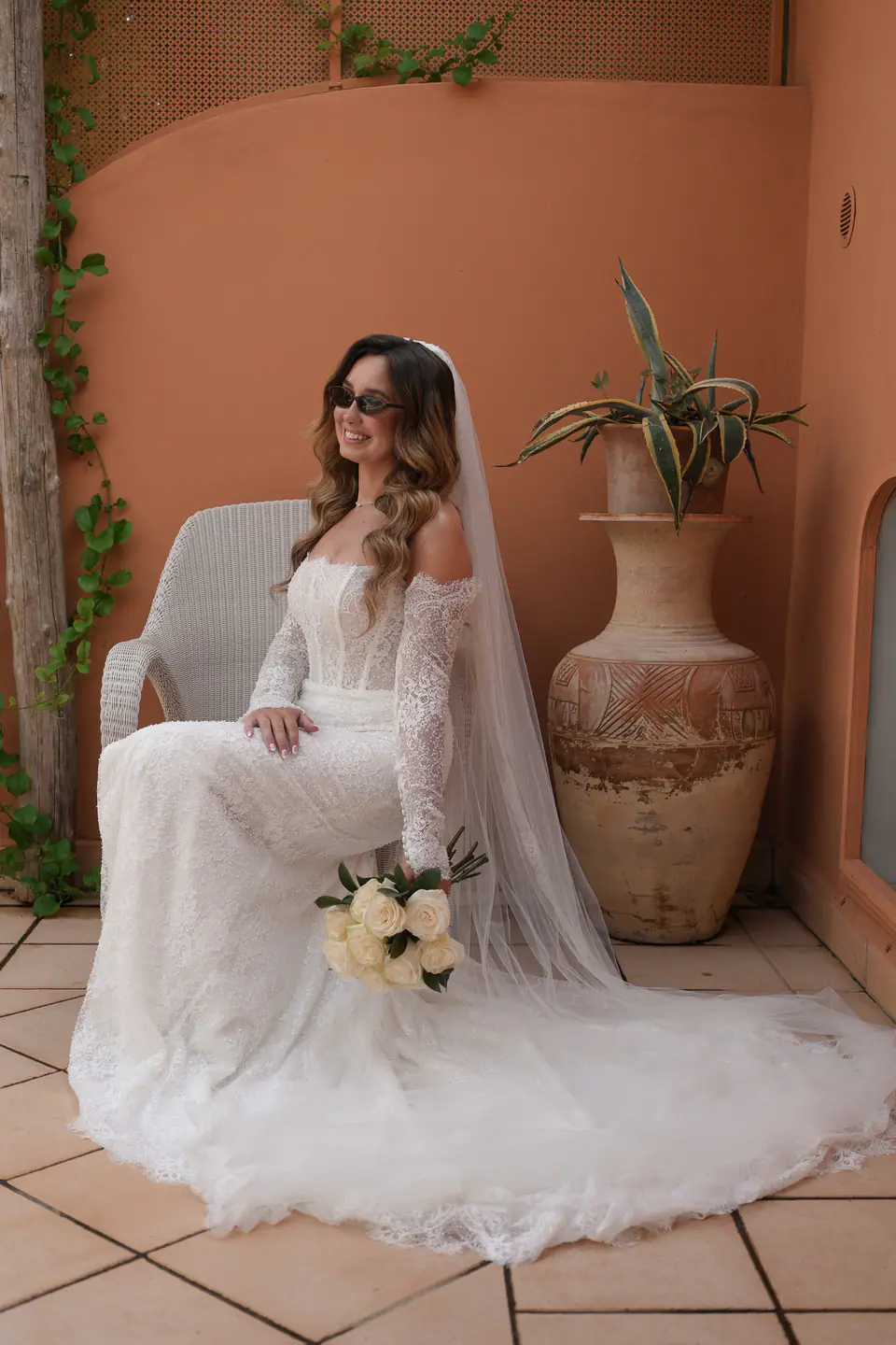 Bride in sunglasses sitting with her bouquet beside a terracotta vase.