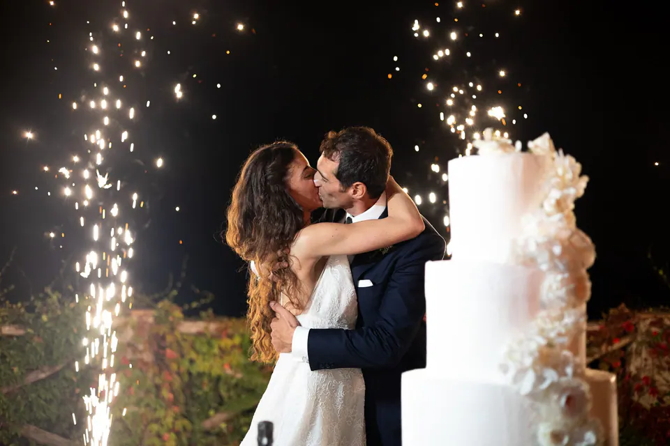 Bride and groom kissing beside the wedding cake as sparklers light up the night.