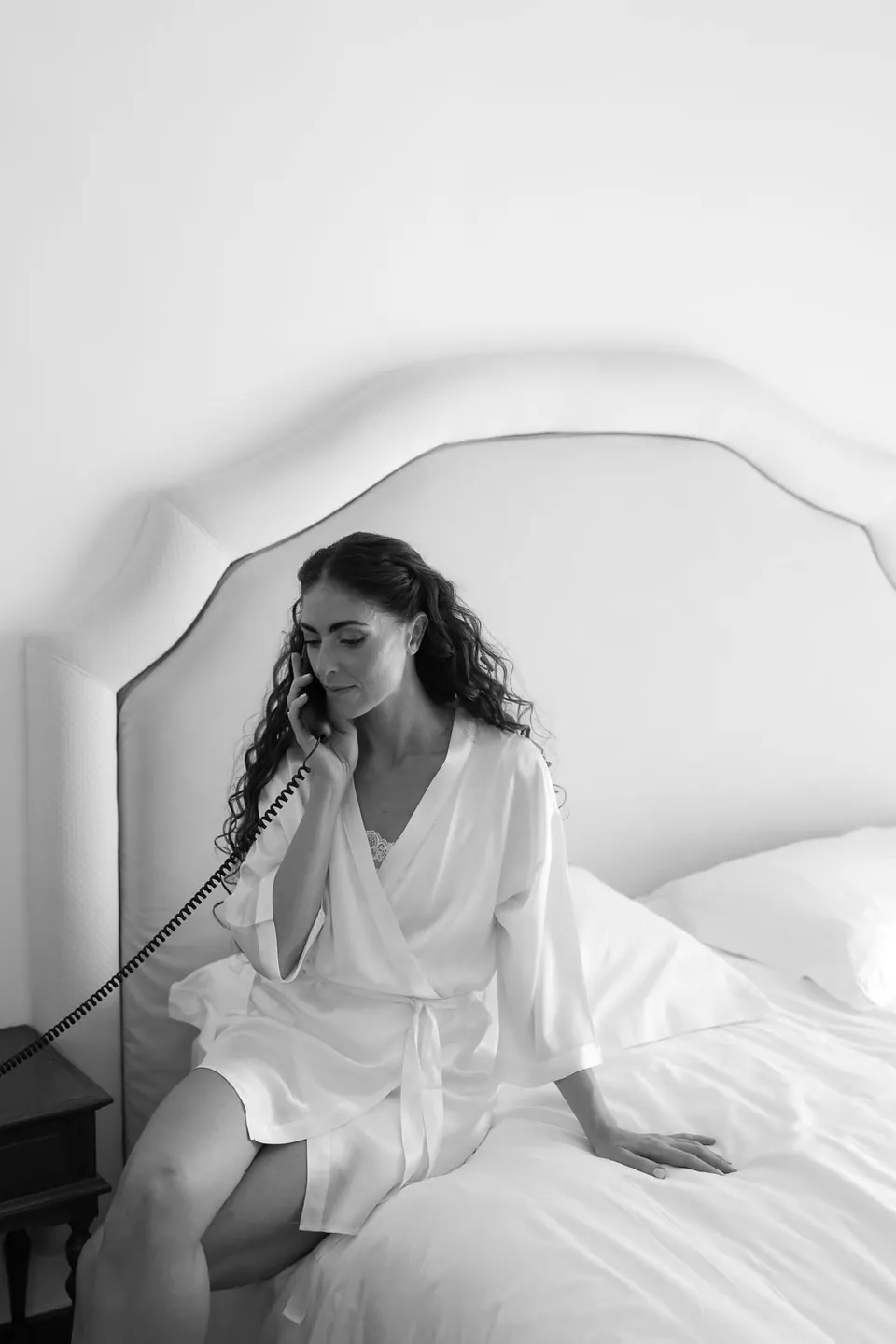 Black and white photo of the bride in her robe sitting on the bed, on the phone.