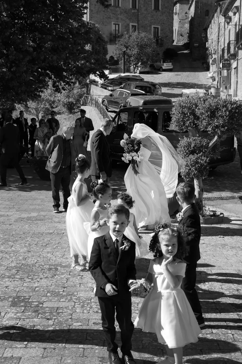 Foto in bianco e nero degli sposi che attraversano il borgo con il velo della sposa che vola.