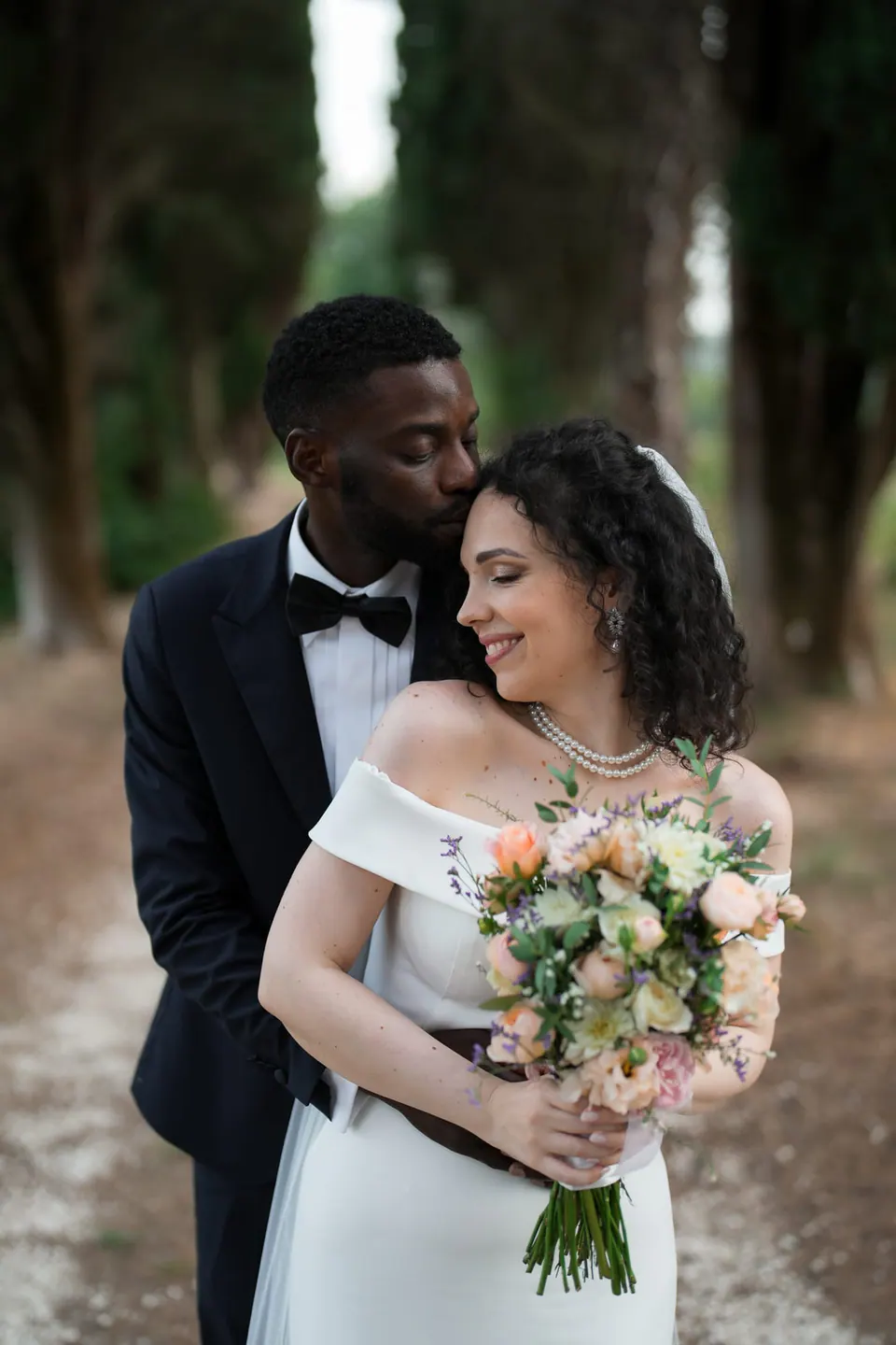 El novio besa la cabeza de la novia, que sostiene su ramo de rosas melocotón y blancas, entre los árboles.