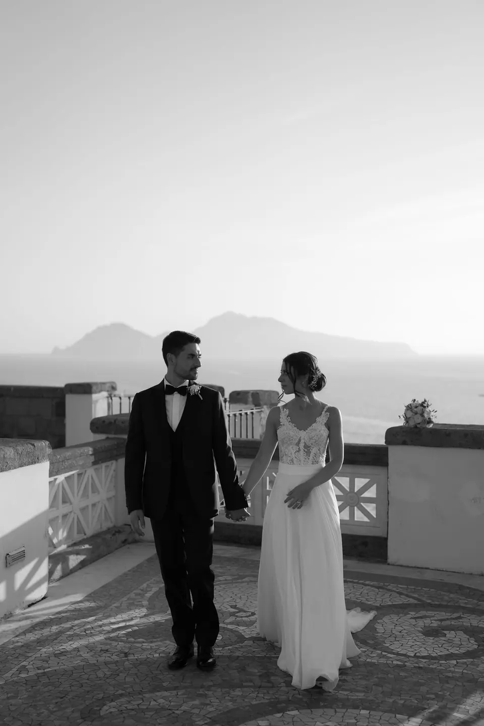 Couple standing together on the terrace with the island of Capri in the distance, poetic coastal moment.