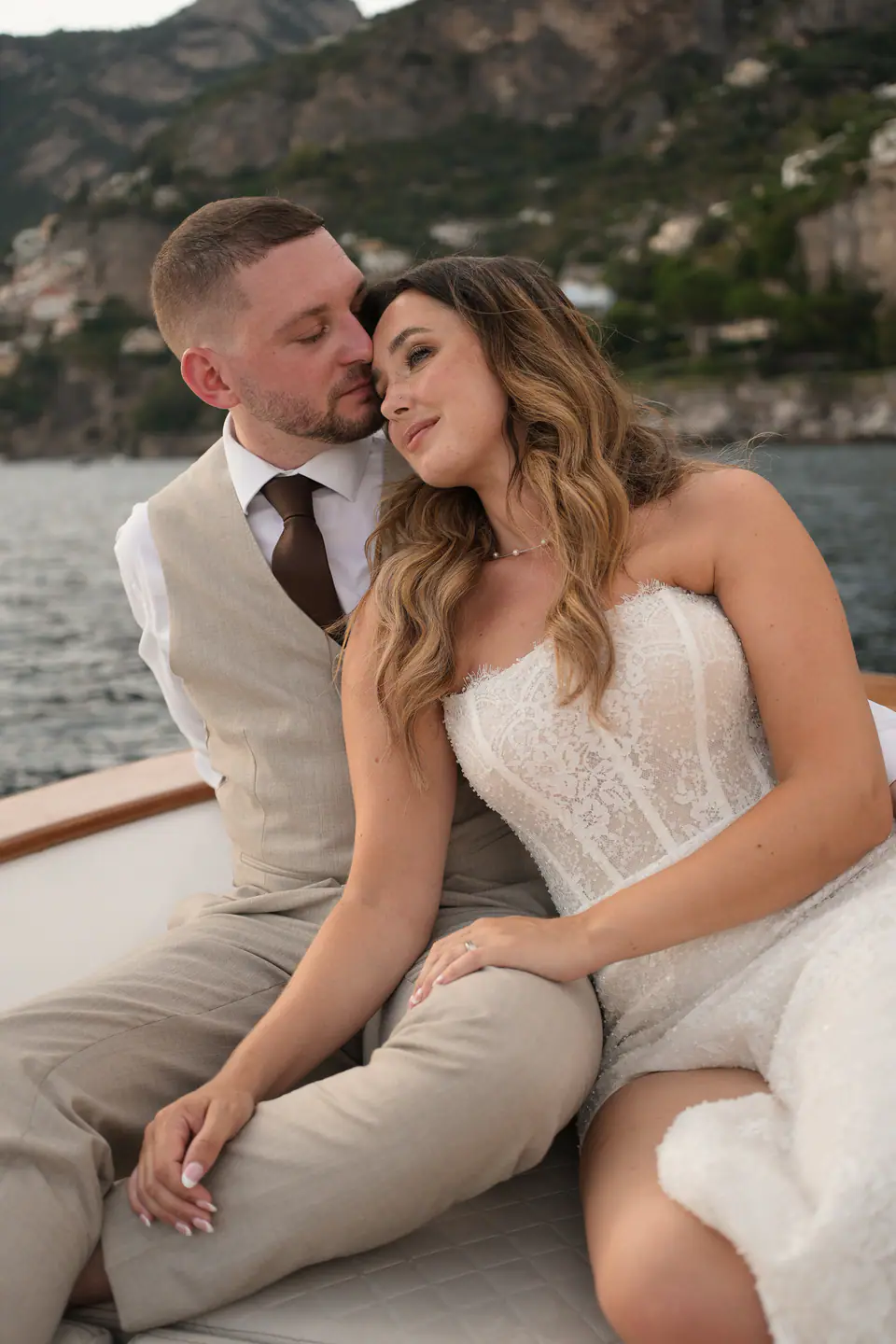 Couple sitting together on the bow of a boat at golden hour, the coast behind them.